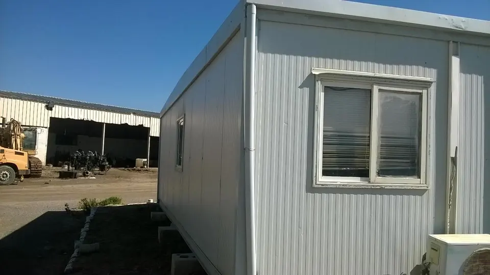 Module de base de vie blanc posé sur des appuis béton, avec climatiseur extérieur, près d'un hangar et d'un engin de chantier