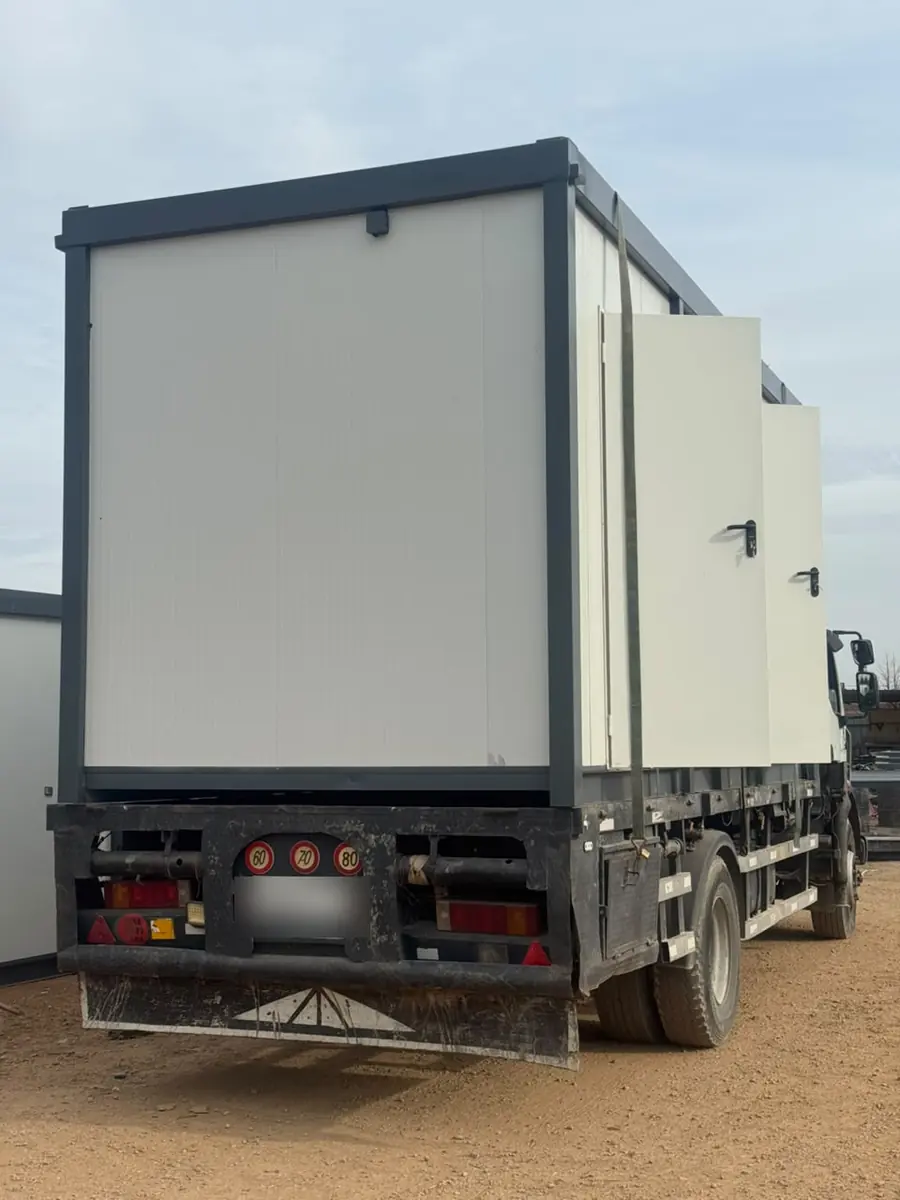 Module préfabriqué blanc chargé sur un camion porteur, vu de l'arrière sur un terrain de terre battue, avant le départ