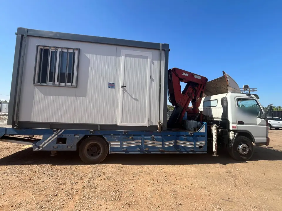 Bungalow de chantier blanc chargé sur le plateau d'un camion équipé d'une grue auxiliaire, sur un terrain en terre